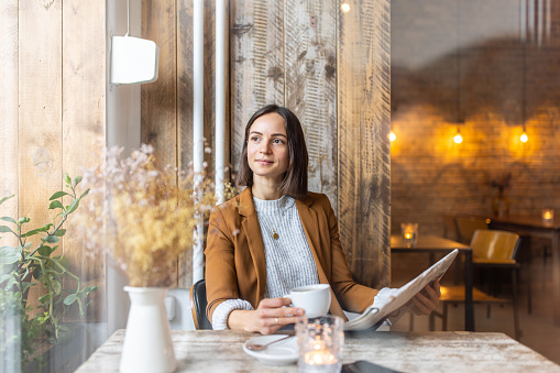 Woman sitting at a table in a coffee shop holding a cup of coffee and newspaper looking outside window. Businesswoman taking a break from work and relaxing in a coffee shop.