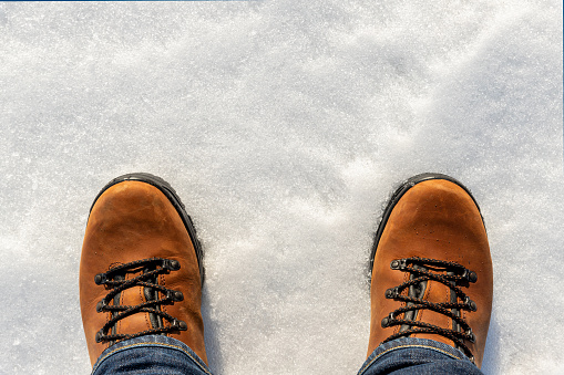 Closeup top above view of male legs in jeans and brown leather warm boots isolated on white icy snow surface background. Detail waterproof shoes. Winter walking and hiking wild nature outdoor concept.