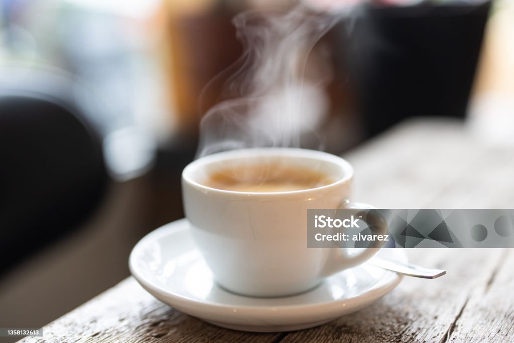 Refreshing hot cup of coffee at a cafe Steam rising from a white cup of hot coffee with a spoon on a saucer over a wooden table in the cafe. Close-up of a refreshing hot cup of a coffee at a cafe. Coffee - Drink Stock Photo