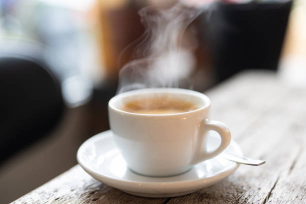 Refreshing hot cup of coffee at a cafe Steam rising from a white cup of hot coffee with a spoon on a saucer over a wooden table in the cafe. Close-up of a refreshing hot cup of a coffee at a cafe. tea hot drink stock pictures, royalty-free photos & images
