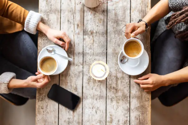 Top view cropped shot of two women sitting at cafe with coffee cups and mobile phone over table. Female friends meeting a coffee shop.