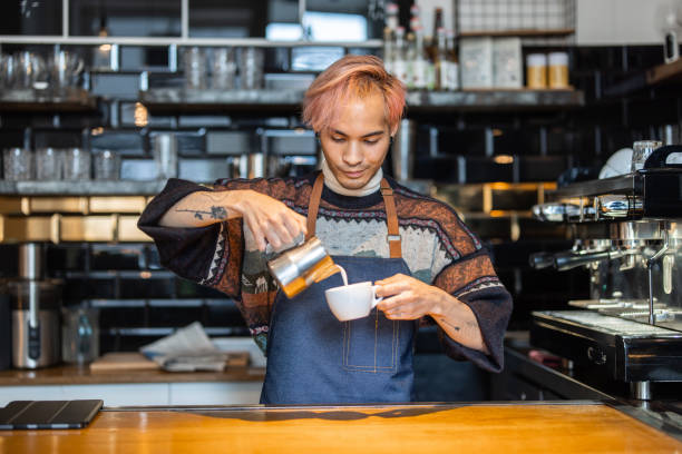 Barista preparing a latte coffee in cafe Man making latte art with frothed milk on coffee shop kitchen. Barista standing behind kitchen counter pouring milk in a coffee latte cup barista stock pictures, royalty-free photos & images