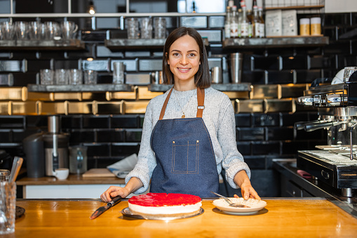 Portrait of a confident young woman cafe worker standing behind kitchen counter with cheese cake. Female cafe owner looking at camera and smiling.