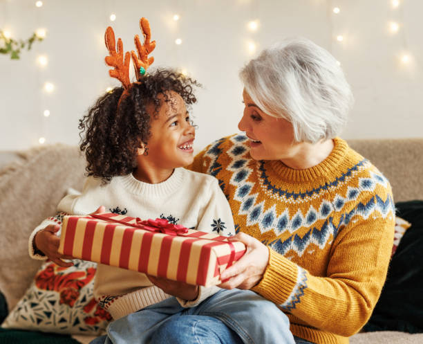 クリスマスプレゼントで祖母の膝の上に座って幸せなアフロアメリカのカーリー少年は、カメラで微笑む - grandmother giving gift child ストックフォトと画像
