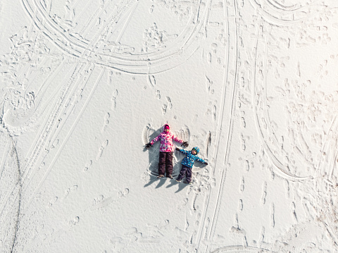 Aerial top view pair two cute little sibling kids boy girl have fun play make snow angel on white frozen lake ice surface landscape bright sunny cold season day. Outdoor family winter activity