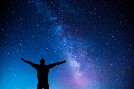 Silhouette of a man with hands raised up, standing, enjoying, loving, embracing the beauty of night sky with milky way.