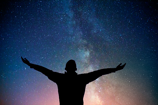 Silhouette of a man with widespread arms, standing, enjoying, loving, embracing the beauty of night sky with milky way.