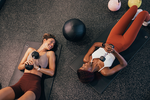 Two motivated fit female friends working out together at the gym, doing exercises on a mat.