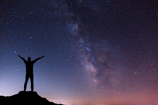 Silhouette of a man standing on the top of the mountain with hands raised up, enjoying, loving, embracing the beauty of night sky with milky way.