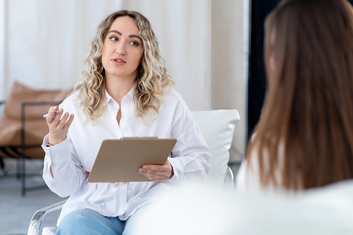 Plus size female psychologist is consulting with unrecognizable patient. Explains carefully and gestures. Holds pen and clipboard.