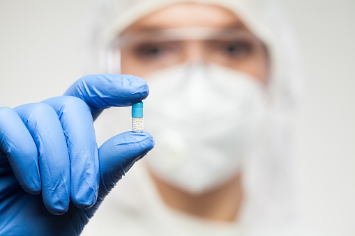 Female UK NHS pharmacist wearing PPE personal protective equipment,coveralls,face mask,eyewear,glove,holding blue pill,selective focus,patient cure remedy for illness disease,prescription drug concept