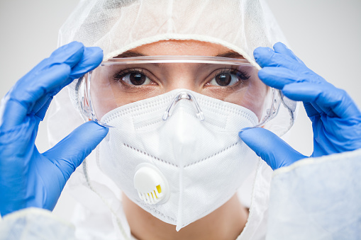 Female NHS doctor,nurse or lab tech virology scientist in PPE,putting on protective eyewear goggles,wearing blue latex gloves and white face mask,closeup headshot portrait isolated on white background