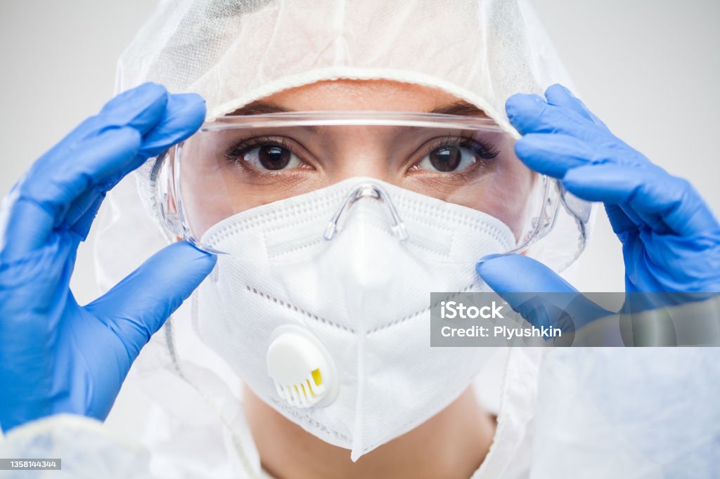 Female NHS doctor,nurse or lab tech virology scientist in PPE,putting on protective eyewear goggles Female NHS doctor,nurse or lab tech virology scientist in PPE,putting on protective eyewear goggles,wearing blue latex gloves and white face mask,closeup headshot portrait isolated on white background N95 Face Mask Stock Photo