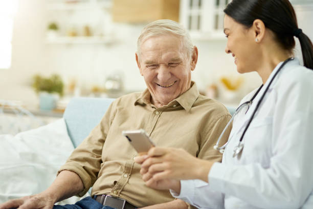istock Smiling nurse and senior citizen sitting on sofa and using phone 1358150755