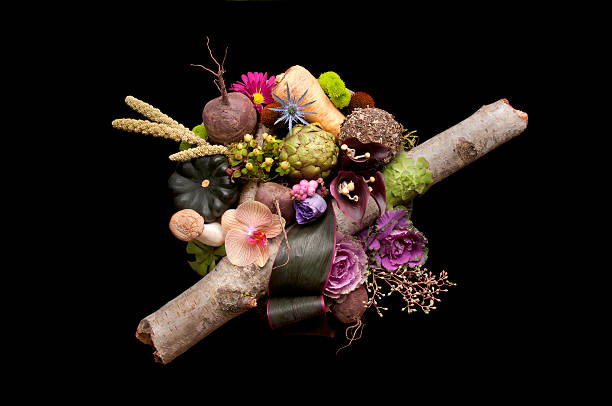 A top-down view of an arrangement of autumn vegetables and flowers in a large bowl against a black background.  Vegetables: 2 types of mushrooms, 3 beets, 2 kale, parsnip, artichoke, and an acorn squash.  Flowers: orchid, chrysanthemum, calla lilies, succulents, eryngium, echinacea, lisianthus.