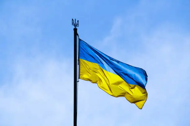 Photo of Ukrainian flag in the rays of the rising sun on a background of sky. Bicolor blue and yellow national flag of Ukraine on a flagpole and coat of arms of Ukraine trident. Official symbol of Ukraine