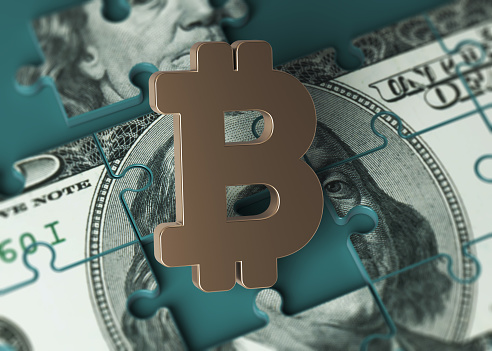 Puzzle-shaped 100 American dollar banknote and gold-colored Bitcoin symbol. On a charcoal green-colored background. Horizontal composition with copy space. Isolated with clipping path.