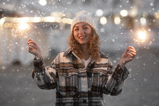 Cozy portrait of beautiful smiling caucasian woman 25-30 years old in fashion coat and knitted hat celebrating new year party at street with sparklers bengal lights on winter holidays outdoor.