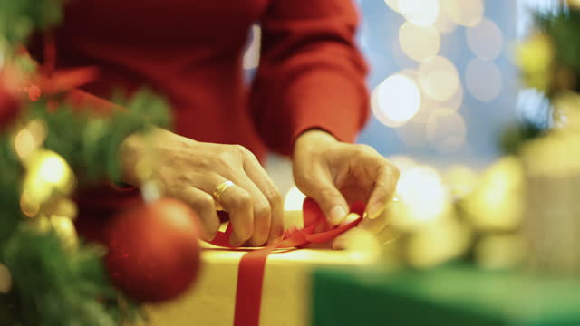 Hands of woman in red winter dress holding and using ribbon to tie bow for a wrapped gift box for Christmas present with Xmas tree in foreground and colorful lights bokeh in blur background.