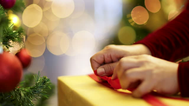 Hands of woman in red dress holding and using ribbon to tie bow for a wrapped gift box for Christmas present with Xmas tree in foreground and colorful lights bokeh in blur background.