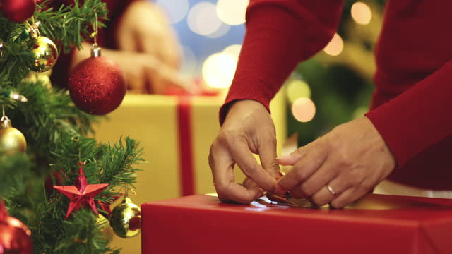 Hands of two women in red dress holding and using ribbon to tie bow for a wrapped gift box for Christmas present with Xmas tree in foreground and colorful lights bokeh in blur background.
