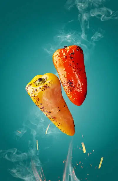 Hot grilled orange and yellow bell peppers hovering over flames in sparks and white haze on turquoise background. Natural food concept
