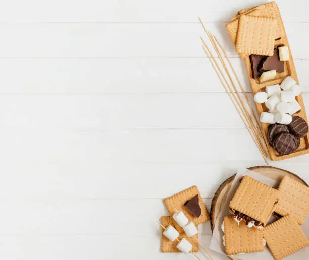 S'mores roasting marshmallows with chocolate between cookies on white wooden background with blank space for text. Top view, flat lay.