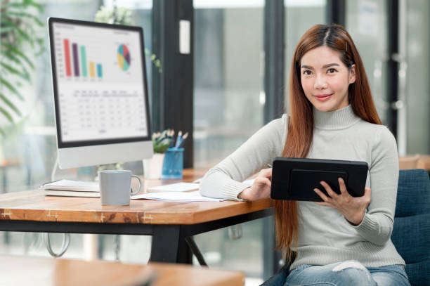 istock Asian woman holding portable tablet, smiling and looking at camera while sitting at office. 1358201218