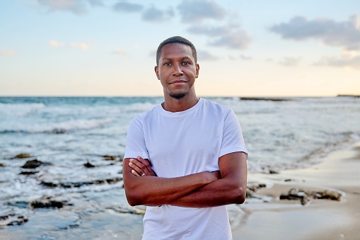 Outdoor portrait of a smiling young African American man looking into the camera. Positive confident 30s man with crossed arms, sky sea beach background. People, lifestyle, health, lifestyle concept