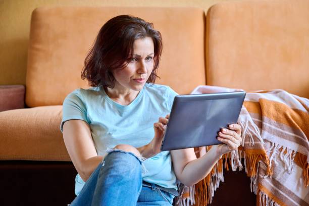 istock Middle aged woman sitting on sofa at home, relaxing using digital tablet 1358202186