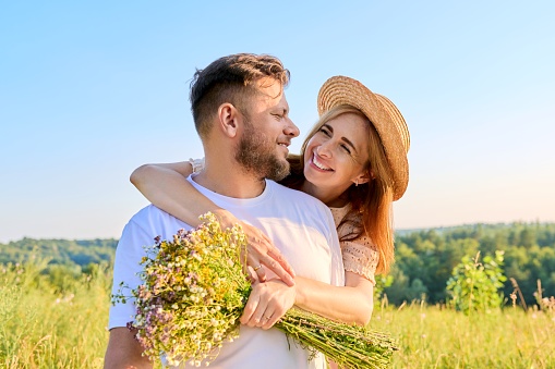 Happy middle-aged couple in love embracing, with bouquet of wildflowers, outdoors on summer nature meadow. Date, anniversary, family, wedding, celebration, relationship, 40s people concept