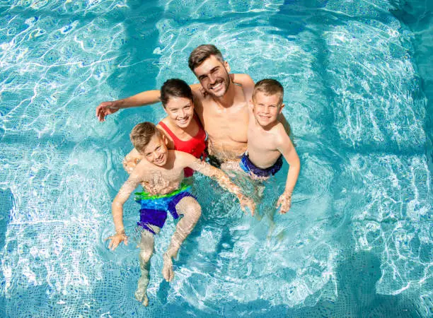 Photo of Top view of smiling family of four having fun and relaxing in indoor swimming pool at hotel resort.