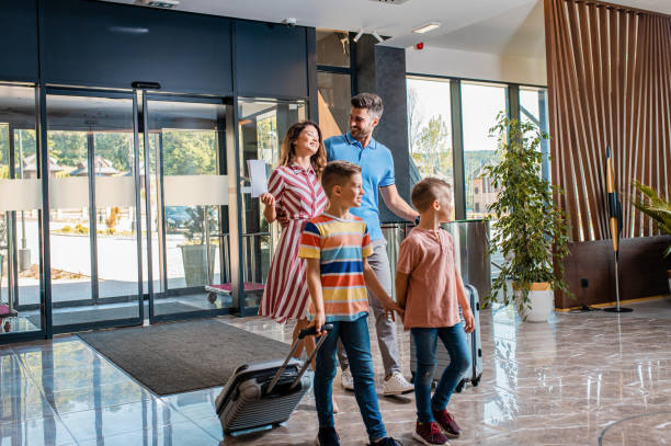 네 명의 웃는 가족이 호텔 로비에 들어와 리셉션에서 휴가를 확인합니다. - 호텔 뉴스 사진 이미지