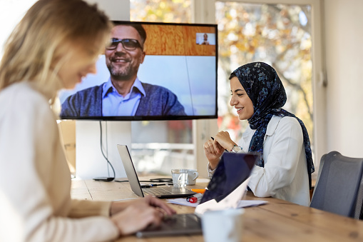 Two businesswomen working at startup office with male colleague connecting on a video conference. Startup team planning in board room with a colleague on video call.