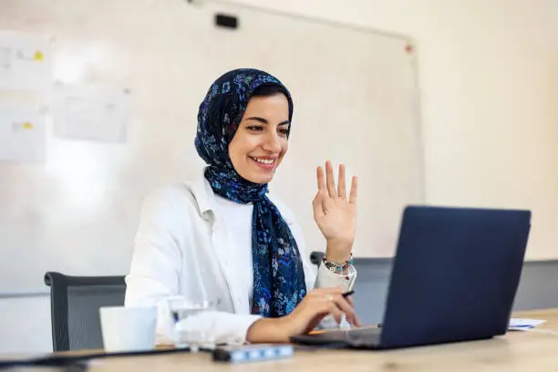 Businesswoman working in the office doing a video call with laptop. Female professional working at a startup and making a video call and waving hand.