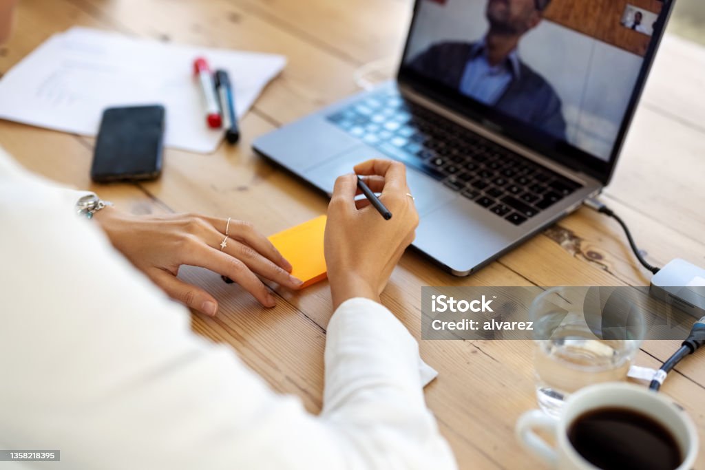 Close-up of a businesswoman having a video conference and taking notes Close-up of a female executive sitting at her office desk making a video call with laptop computer. Businesswoman having a video conference and taking notes. Web Conference Stock Photo