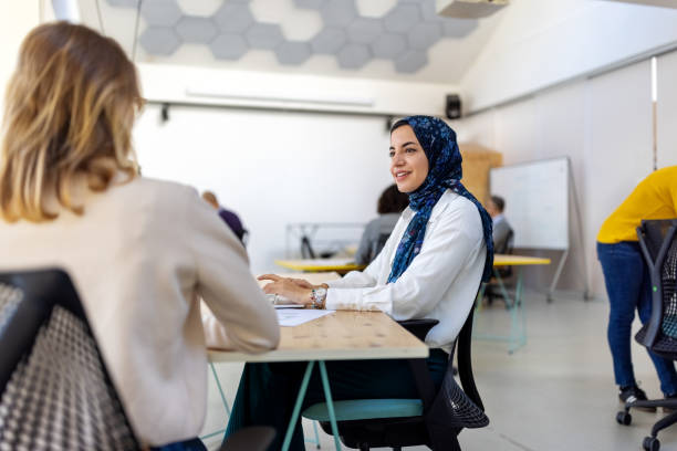 Middle eastern young woman talking with a female colleague working alongside her at startup office. Female startup professionals working together at coworking space.