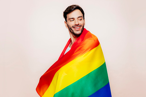 Side view of cheerful homosexual man with a gay pride flag at studio over beige background. Lgbtq flag, rainbow flag, celebrating parade.