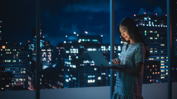 istock Financial Analyst Using Laptop Computer, Standing Next to Window with Beautiful Night City Skyline with Skyscrapers. Confident Asian Businesswoman Working in Modern Corporate Office. 1358221033