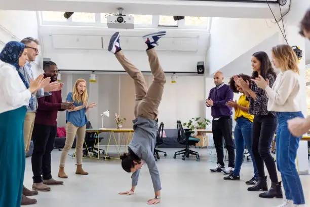 Young man doing a handstand with the team standing by and cheering at startup office. Office staff celebrating success at the office.