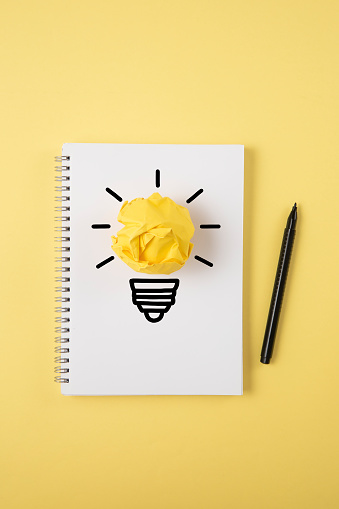 Light bulb made of yellow paper on white notepad on yellow background.