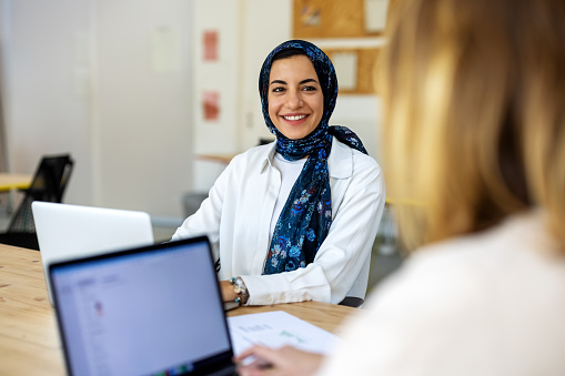 Smiling businesswoman wearing headscarf talking with a female colleague working on laptop at startup office.  Middle eastern woman sitting at coworking desk discussing with a colleague.
