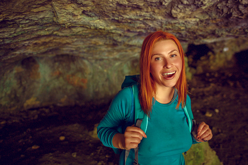 Laughing and cheerful redhead woman in mountain cave holding backpack