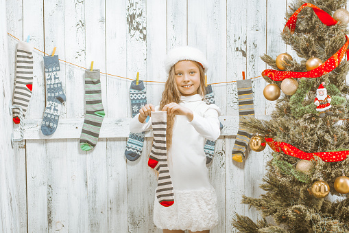 Beautiful Caucasian Girl of 9s in a Delicate White Dress and a Winter Hat Holds a Striped Christmas Sock for Gifts next to a Decorated Green Christmas Tree with Golden Balls and Red Ribbon. Christmas Stocking . Close-up. High quality photo