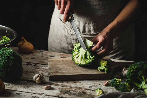 Woman cook cutting a slice of fresh broccoli in a kitchen on top of a wooden cutting board