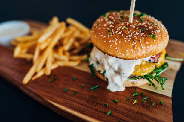 Close-up of a burger with a wooden spike served on a wooden board together with French fries. Soft bun, burger sauce, crispy salad, perfect piece of meat and melted cheese - the perfect combination of taste and texture. bacon cheeseburger stock pictures, royalty-free photos & images