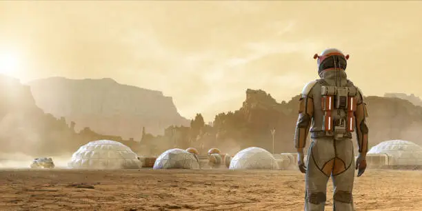 Rear view of an astronaut on Mars looking at the base camp settlement built near a rocky mountain range. The settlement consists of domes and tunnels linked together. A Mars Rover vehicle is driving in the distance.