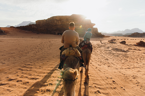 Three mixed-raced explorers of woman and men having camel riding through the majestic arid desert landscape during bright sunset in the Middle East