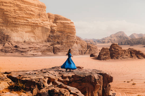 Woman in blue dress contemplating the scenic landscape of Wadi Rum desert Beautiful female in dress walking at the edge of the cliff enjoying the Martian world with red sands and the mountains in Jordan jordan middle east photos stock pictures, royalty-free photos & images