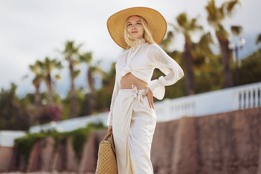 Stunning blonde woman in summer beach outfit relaxing outdoors against sea resort and palm trees on the background. A fashionable romantic young adult lady wearing a trendy vintage straw hat, white blouse, and skirt, standing at the sea coast